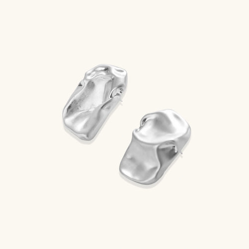 Pair of sculptural silver ear cuffs with organic fluid shapes and polished finish against cream background.