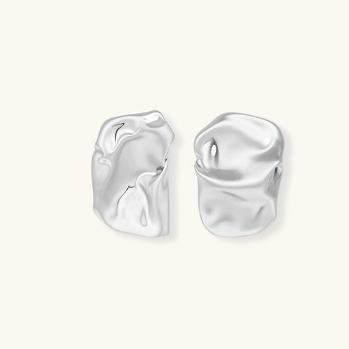 Pair of sculptural silver ear cuffs with organic fluid shapes and polished finish against cream background.